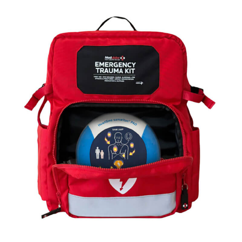 A red Aero Healthcare Modulator Trauma Backpack M600B with a partially open front pocket reveals a defibrillator inside. It features an emergency trauma kit label, reflective strip, and meets ANSI Class B First Aid Kit standards.