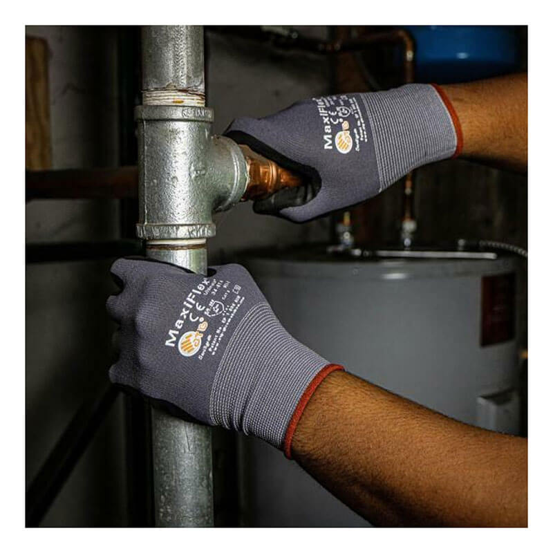 A person wearing PIP ATG G-Tek MaxiFlex Ultimate Nitrile-Coated Gloves 34-874 tightens a metal pipe fitting indoors, with plumbing and a water heater visible in the background.