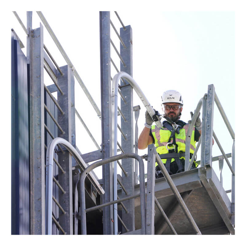 A construction worker in a helmet, safety glasses, and reflective vest stands on scaffolding equipped with the Frontline Securail Pro Vertical Lifeline System VLRS by Frontline Fall Protection, holding the railing and looking downward.