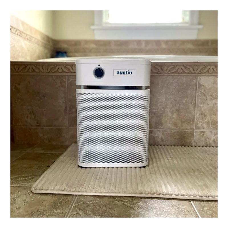 An Austin Air Systems HealthMate Junior Machine A200, featuring a sleek white design, sits on a beige textured rug in a tiled bathroom. Positioned in front of a bathtub with decorative trim and a window in the background, this medical-grade HEPA air purifier seamlessly complements the tranquil space while providing outstanding air quality.