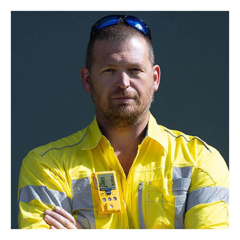 A man in a bright yellow safety jacket with reflective stripes stands with arms crossed, wearing the WatchGas SST4-MINI Diffusion 4-Gas Detector by WatchGas clipped to his chest. He has short hair, a beard, and sunglasses on his head.
