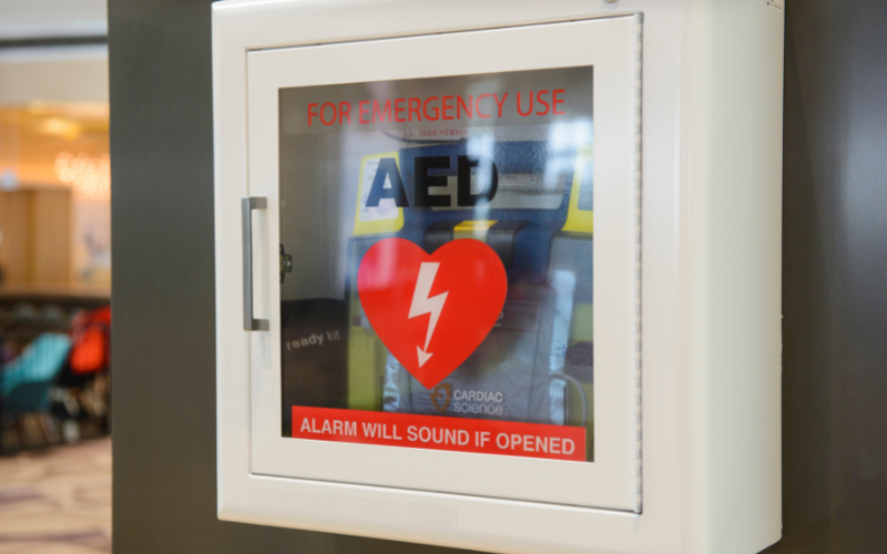 Wall-mounted AED cabinet with transparent door. Visible text "For Emergency Use" and heart symbol with lightning bolt. Alarm warning below. Indoor setting.