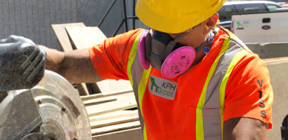 A construction worker wearing a yellow hard hat, orange safety vest, and a respirator mask operates a power saw. They're focused on their task, surrounded by construction materials and a parked truck in the background.