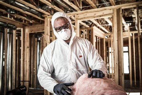 A person wearing protective gear, including a white suit, gloves, goggles, and a mask, handles insulation material inside a building under construction with exposed wooden framing and pipes.
