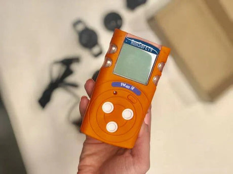 Hand holding orange gas detector with digital display. 