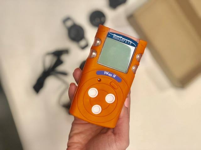 A hand holds an orange gas detection device with a screen and buttons. Other parts and a box are blurred in the background.