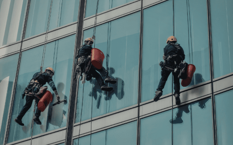 Window cleaners in harnesses and helmets climbing down a high rise building exterior with red buckets
