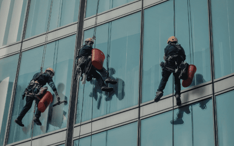 Window cleaners in harnesses and helmets climbing down a high rise building exterior with red buckets