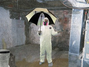 Man wearing hazmat suit and respirator standing in basement area holding umbrella.
