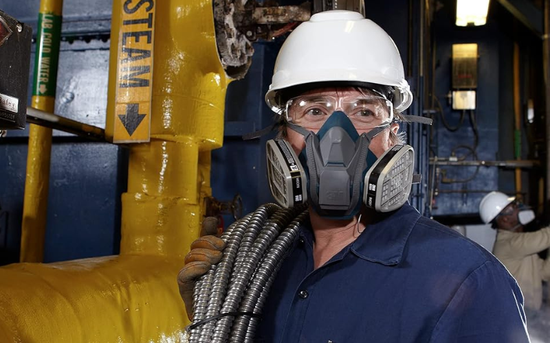 Worker wears helmet, glasses and respirator near pipes of a facility.
