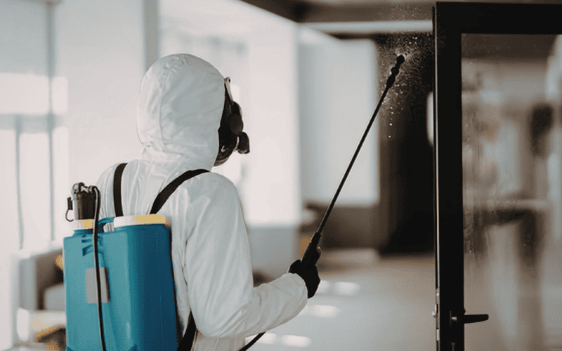 Worker in hazmat suit spraying door with sanitizing solution.