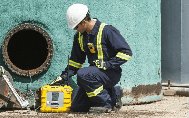 Worker kneels outside of confined space entry holding gas detection device.