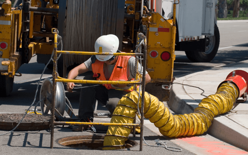 Worker wearing hard hat neat manhole with ventilation going into it.