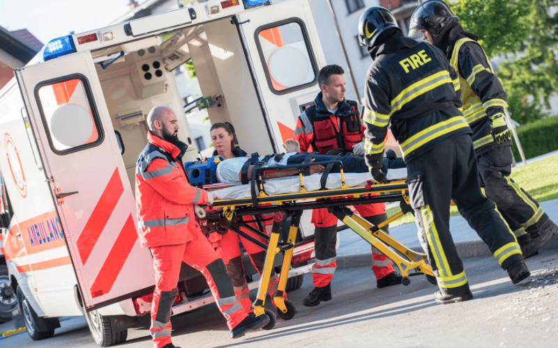 First responders and firefighters push victim on stretcher with ambulance behind them.