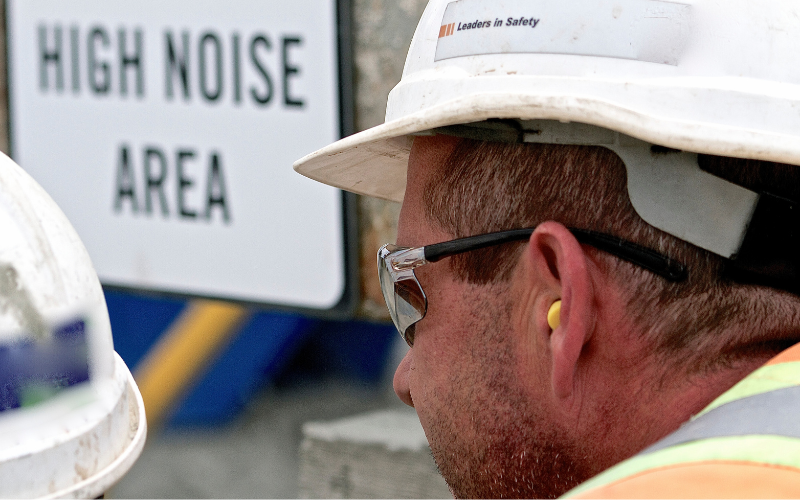 Worker wearing ear plugs with sign in background that reads "High Noise Area"