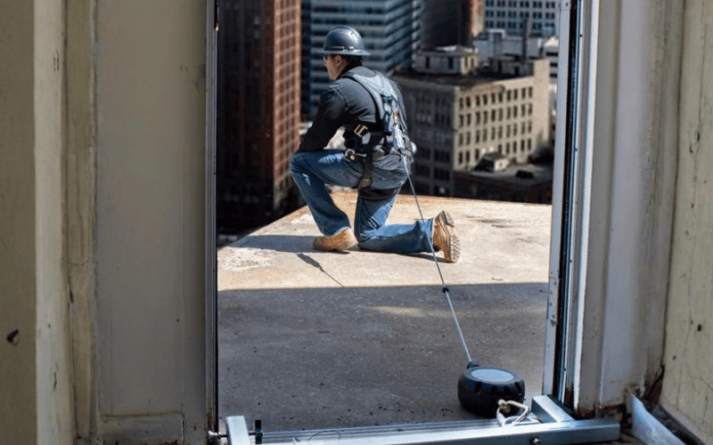 Person on a rooftop wearing heard hat and harness connected to anchorage and line with a cityscape in the background
