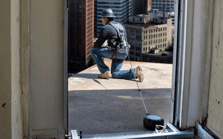 Person on a rooftop wearing heard hat and harness connected to anchorage and line with a cityscape in the background