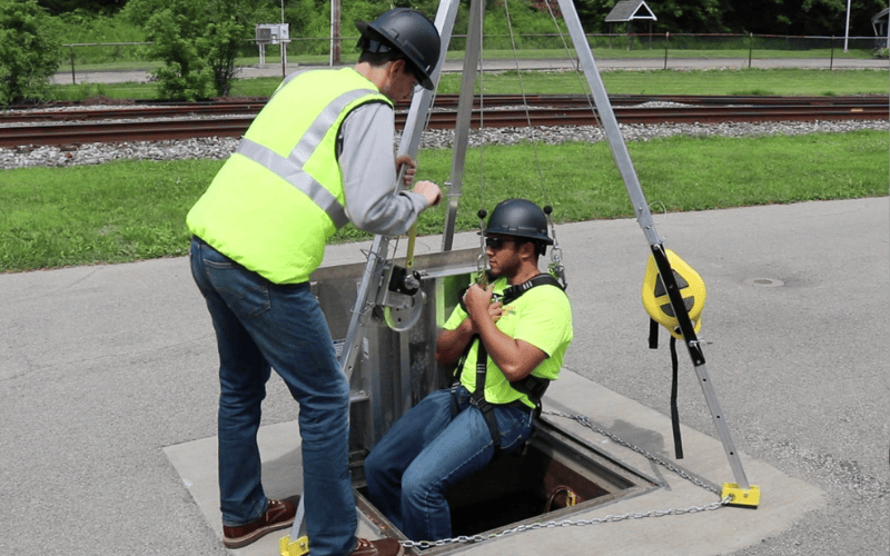 Worker helps other being lowered into a manhole using a tripod.