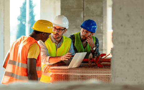 Three construction workers in hard hats and safety vests discussing plans on a laptop at a construction site.