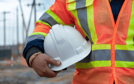 A person wearing a neon safety vest holds a white hard hat. The background is blurred, highlighting construction or worksite safety.
