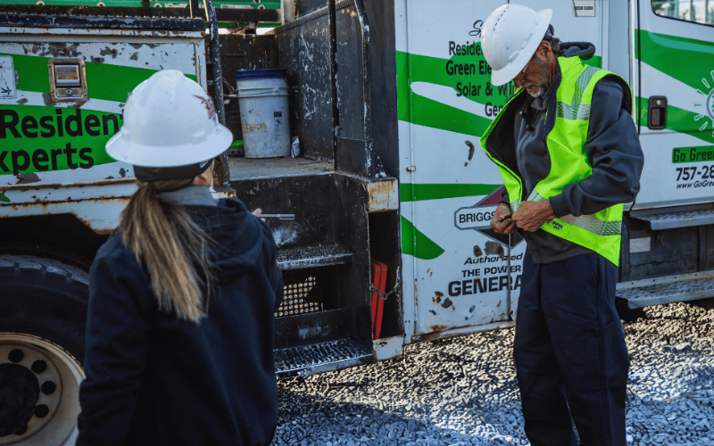 Man putting on FR hi-vis vest while standing next to truck wearing a hard hat. Woman stands nearby wearing hard hat.