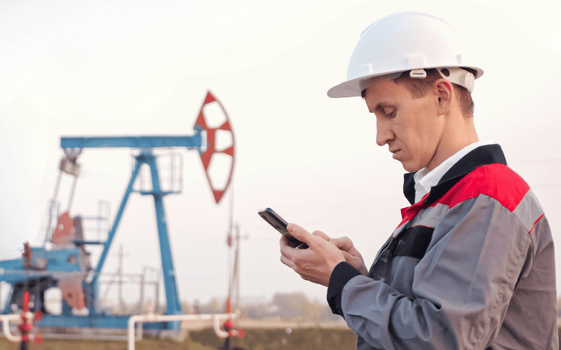 Worker wearing helmet looking down at phone with oil rigs in the distance.