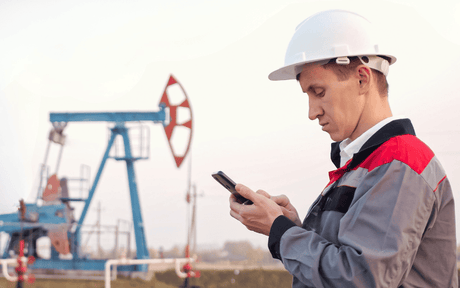 Worker wearing helmet looking down at phone with oil rigs in the distance.