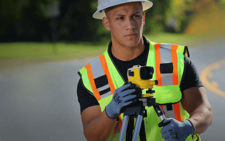 Person in high-visibility vest and hard hat holding a surveying instrument on a road.