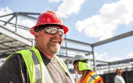 Construction worker wearing a red hard hat, safety glasses with mirrored lens, and reflective vest on a construction site.