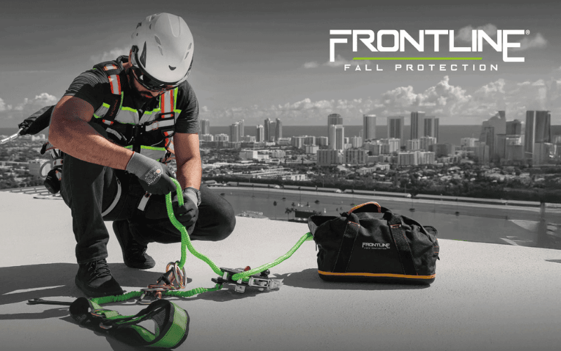 Worker wearing helmet, harness and other fall protection while kneeling on the roof of a building with a skyline behind him.