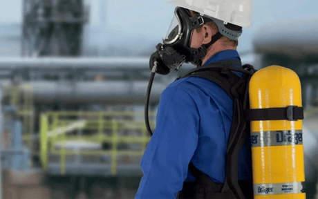 Worker wearing SCBA with yellow tank, helmet and full-face respirator looking towards an industrial plant.