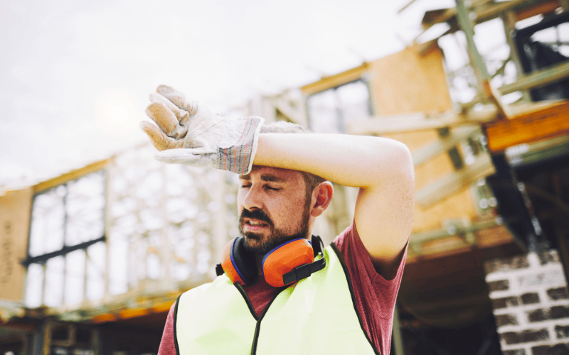 Tired construction worker standing on construction site, wiping sweat from head.