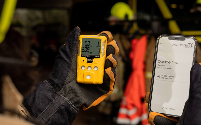 A person in work gloves holds a yellow WatchGas SST4 multi-gas detector in one hand and a smartphone showing the WatchGas device link app in the other.