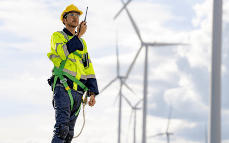 Worker talking to device while wearing hard hat and harness near wind tourbines.