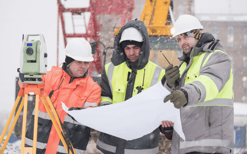 Civil engineers dressed in winter PPE looking at paper at job site where snow is falling down.