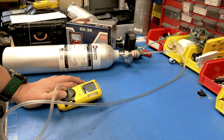 Hand holds gas monitor with tube coming from gas cylinder on blue work desk.