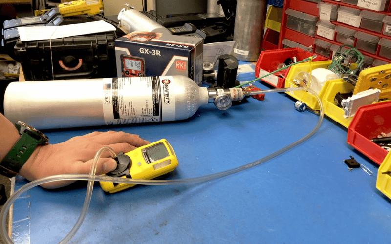 Worker's hand holding a yellow gas monitor at a work desk with calibration gas in the background.