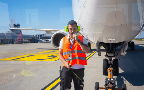 Airport ground crew member in an orange vest stands in front of an airplane, guiding it with a handheld device. Bright, clear day on the tarmac.