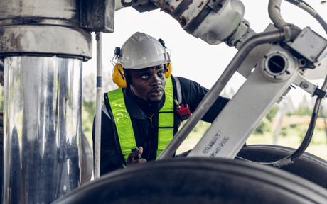 A worker in a white hard hat and safety vest inspects machinery. He wears yellow earmuffs, appearing focused and attentive in an industrial setting.