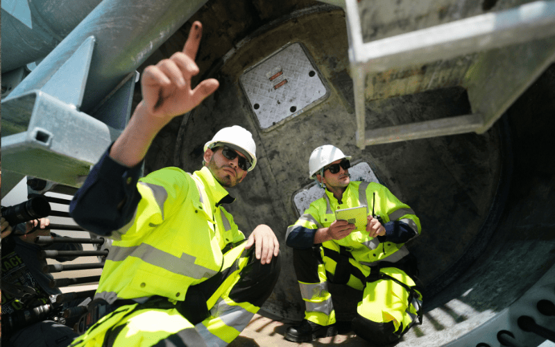 Construction workers in high-visibility gear inspect large industrial machinery.
