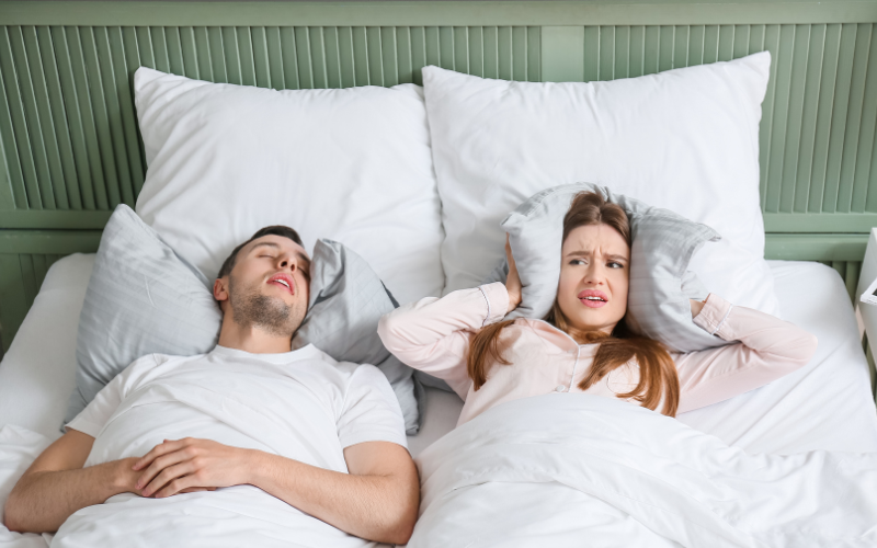 Woman covering ears with pillow while man sleeping next to her snores.