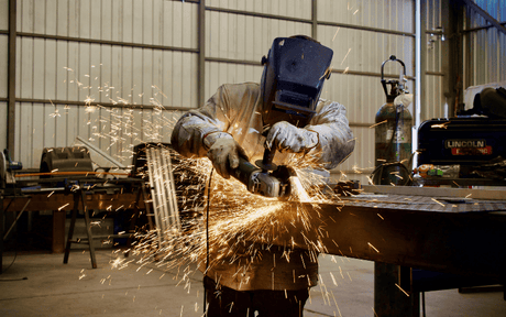 A person wearing a welding helmet and protective gear uses an angle grinder on metal, producing sparks, inside a large industrial workshop.