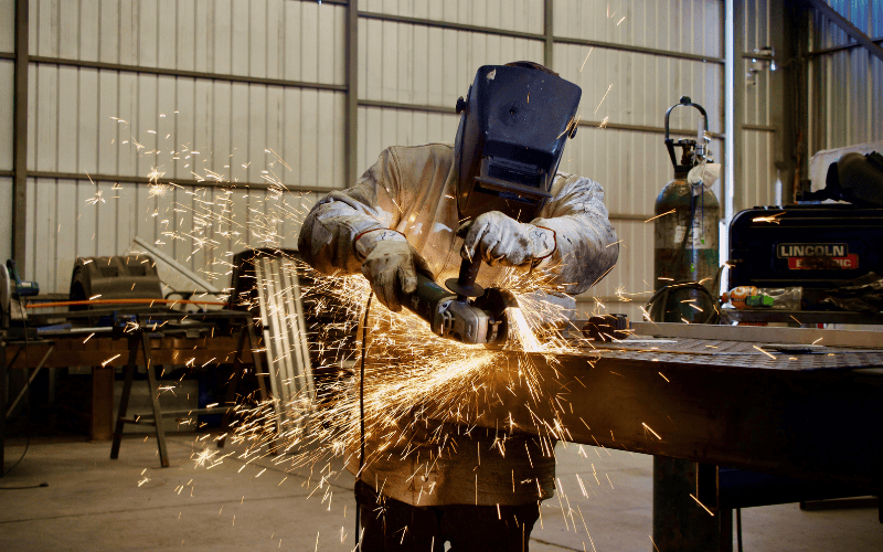 A person wearing a welding helmet and protective gear uses an angle grinder on metal, producing sparks, inside a large industrial workshop.