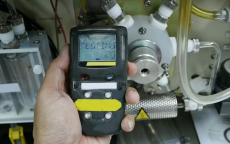 Worker holds up a gas monitor with a machine in the background.