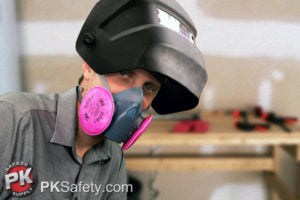 A person wearing a protective welding mask and a respirator with pink filters is standing in a workshop. Tools and wooden frames are visible in the background.