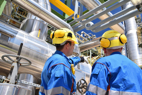 Two industrial workers in blue uniforms and yellow helmets discuss machinery in a factory, with pipes and valves visible. Daylight, professional tone.