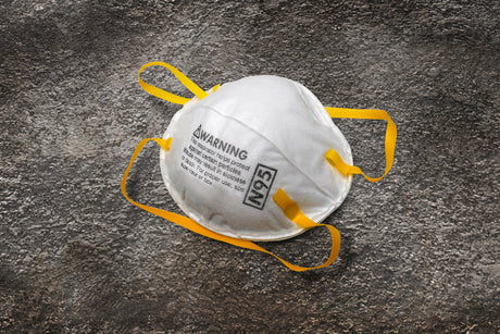 A white N95 mask with yellow straps lies on a textured, gray surface. The mask has printed text and an N95 label, conveying safety and protection.