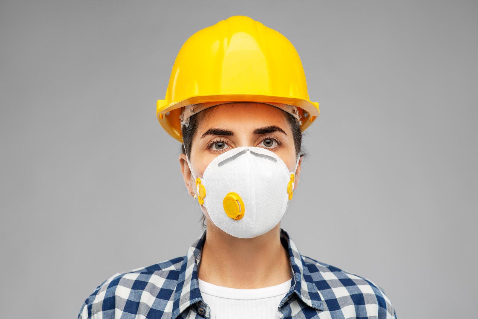 Person wearing a yellow hard hat and white face mask stands against a gray background. They wear a blue plaid shirt, conveying a safety-focused context.