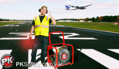 Airline worker standing on runway giving thumbs up. Stands next to ventilator with aircraft flying behind him.