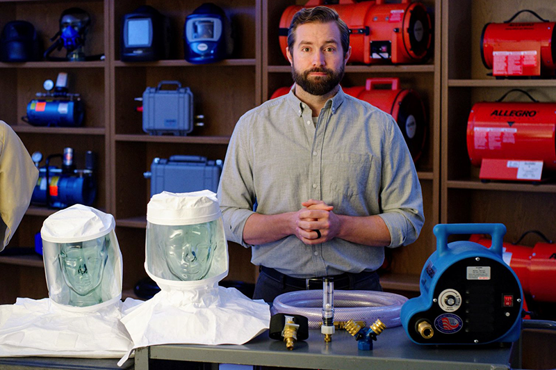 A man stands behind a table displaying protective hoods, hoses, and equipment. Shelves behind him hold red and blue industrial devices. He faces forward with hands clasped, wearing a light gray shirt.
